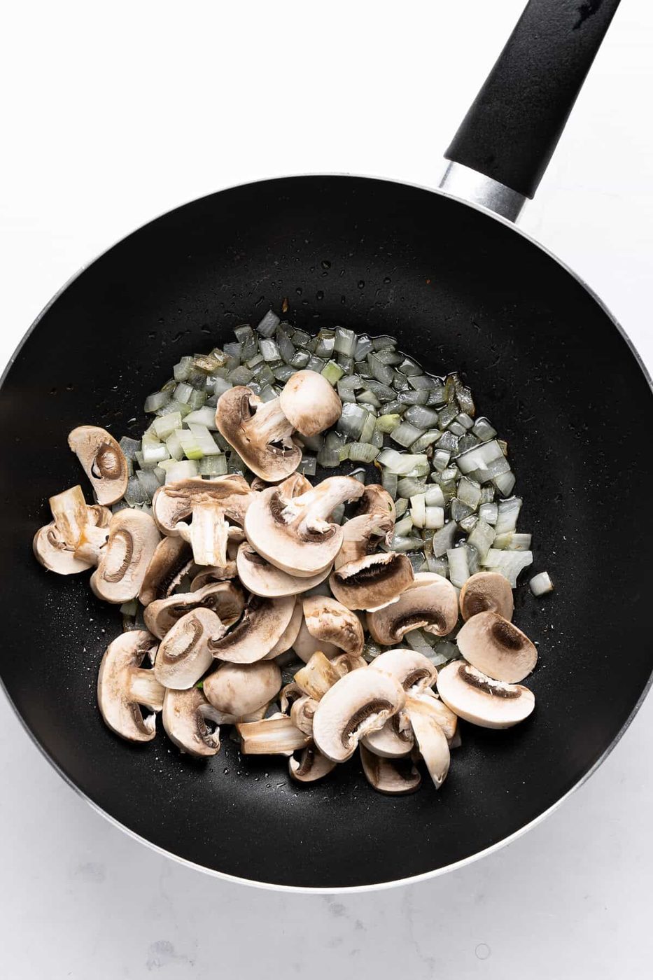 Chopped onions and sliced mushrooms cooking in a black frying pan on a light surface.