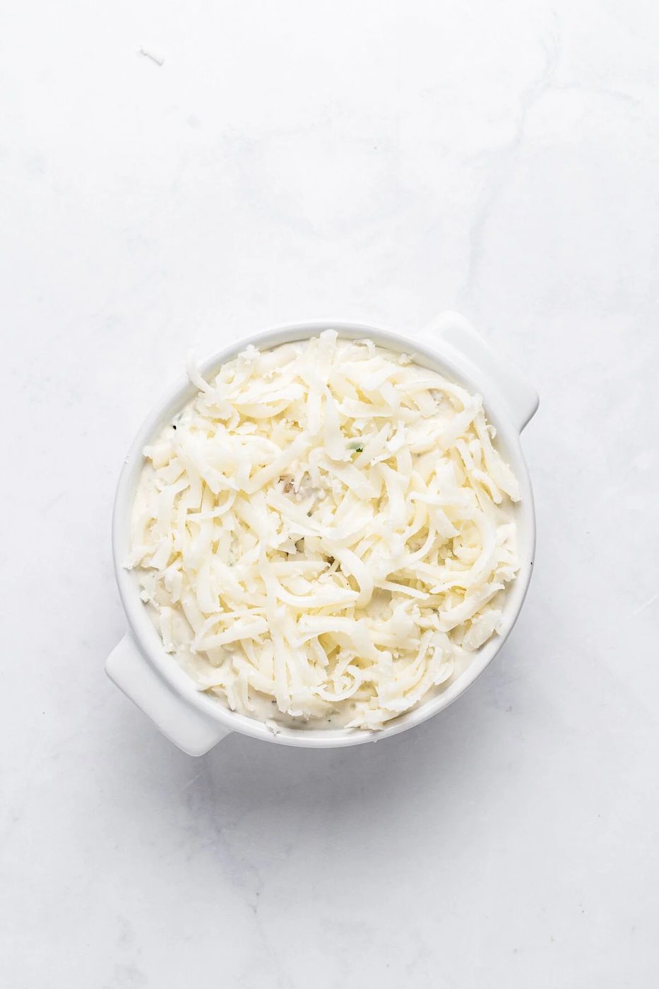 A white ceramic bowl filled with shredded mozzarella cheese placed on a white marble surface.
