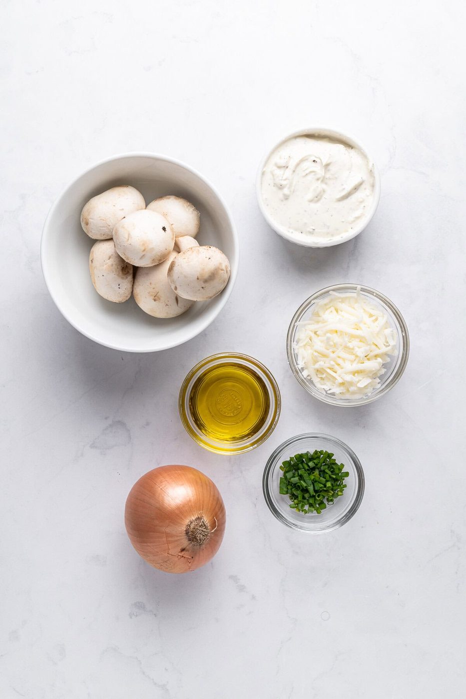 A collection of cooking ingredients on a white surface: mushrooms in a bowl, a bowl of cream cheese, shredded cheese, a small bowl of chopped herbs, a whole onion, and a small bowl of olive oil.