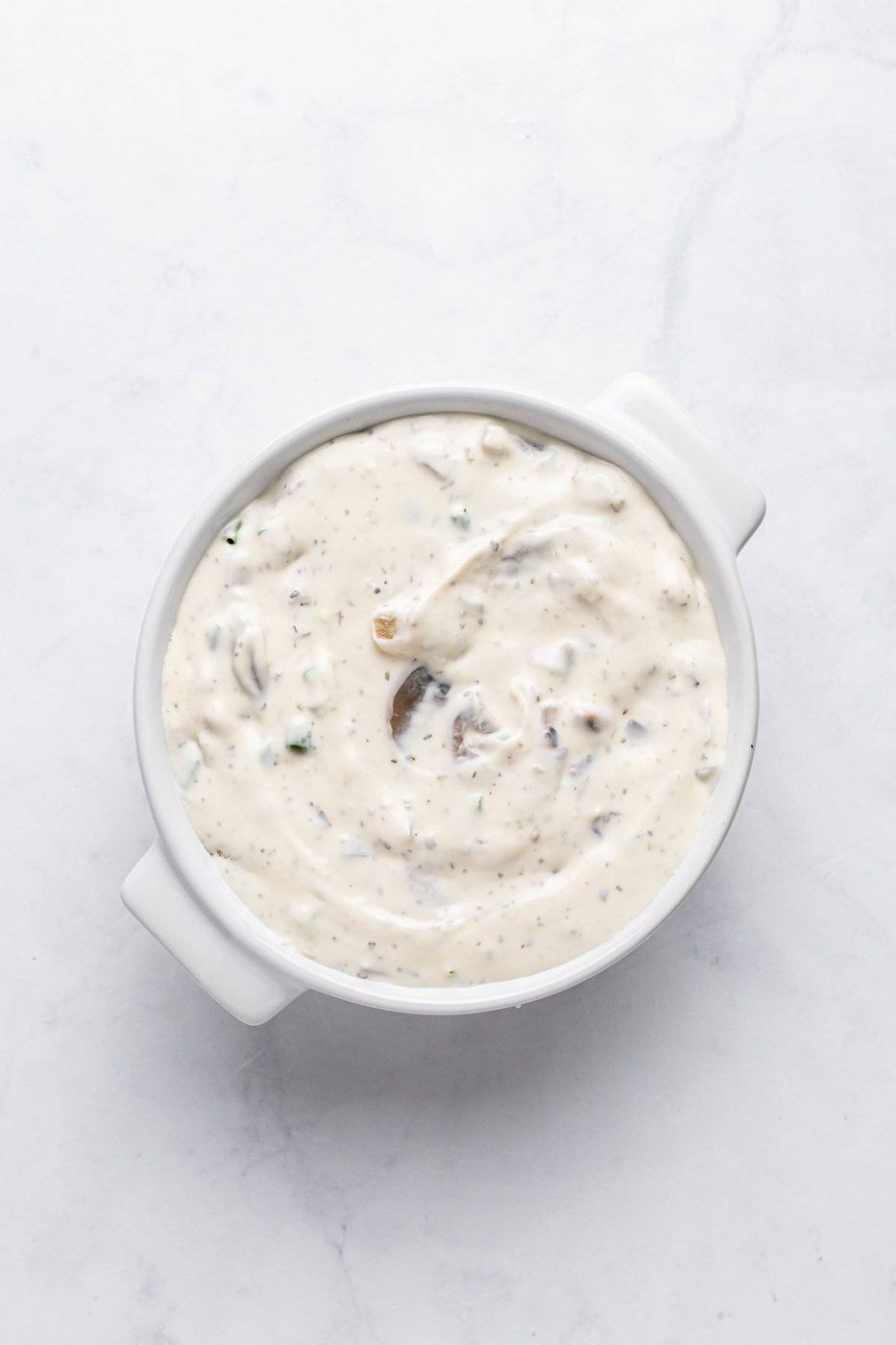 A white ceramic bowl filled with creamy ranch dressing, flecked with herbs, on a marble countertop.