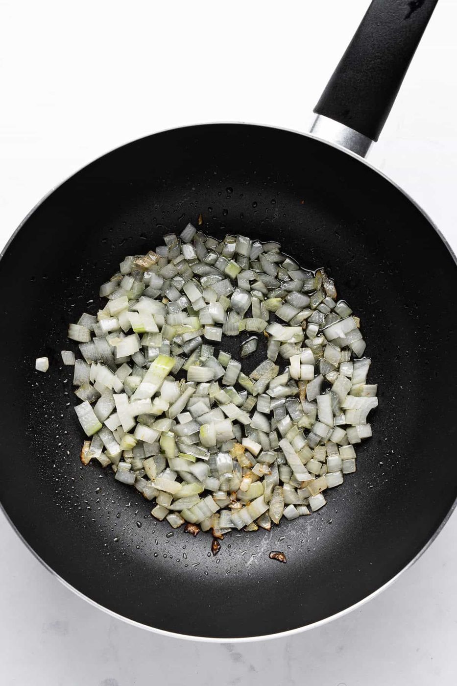 Chopped onions sautéing in a black frying pan on a white surface. The onions are lightly browned, indicating they are being cooked.