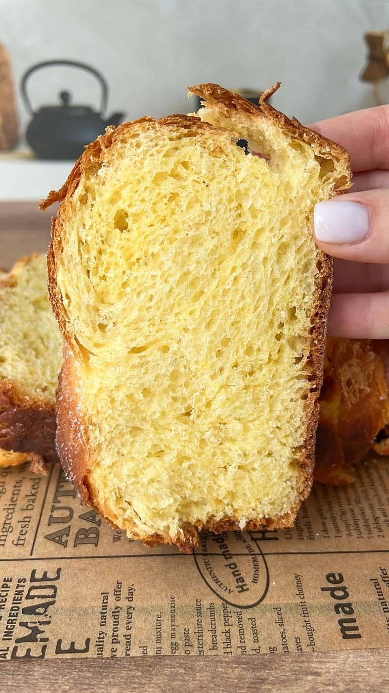 A hand holding a thick slice of golden, fluffy brioche bread with a soft, airy texture. A loaf and more slices are visible in the background on a rustic paper-lined wooden surface.
