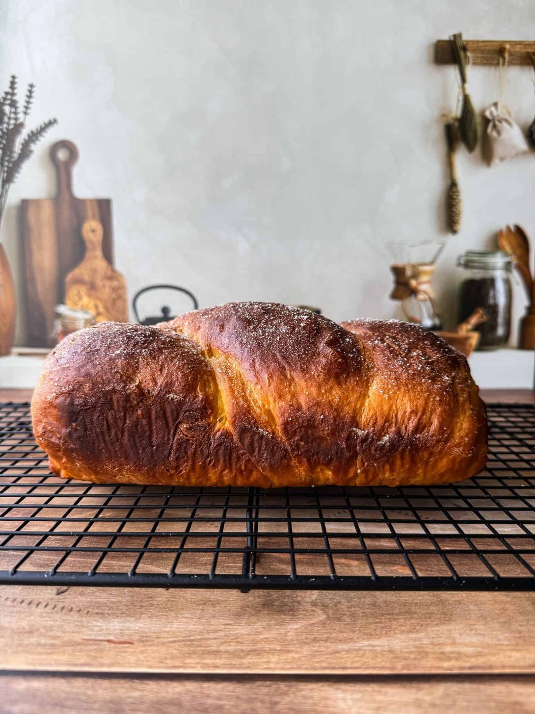 A golden-brown loaf of bread with a sugar-dusted top cools on a black wire rack in a cozy kitchen setting with wooden utensils and jars in the background.