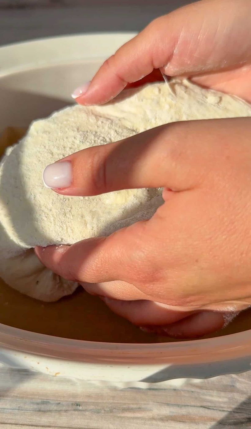 Close-up of hands placing the No Knead Feta Bread in to a sunlit bowl. The focus is on the white flour-dusted dough being worked with care. The hands have manicured nails, and the scene suggests baking in progress.