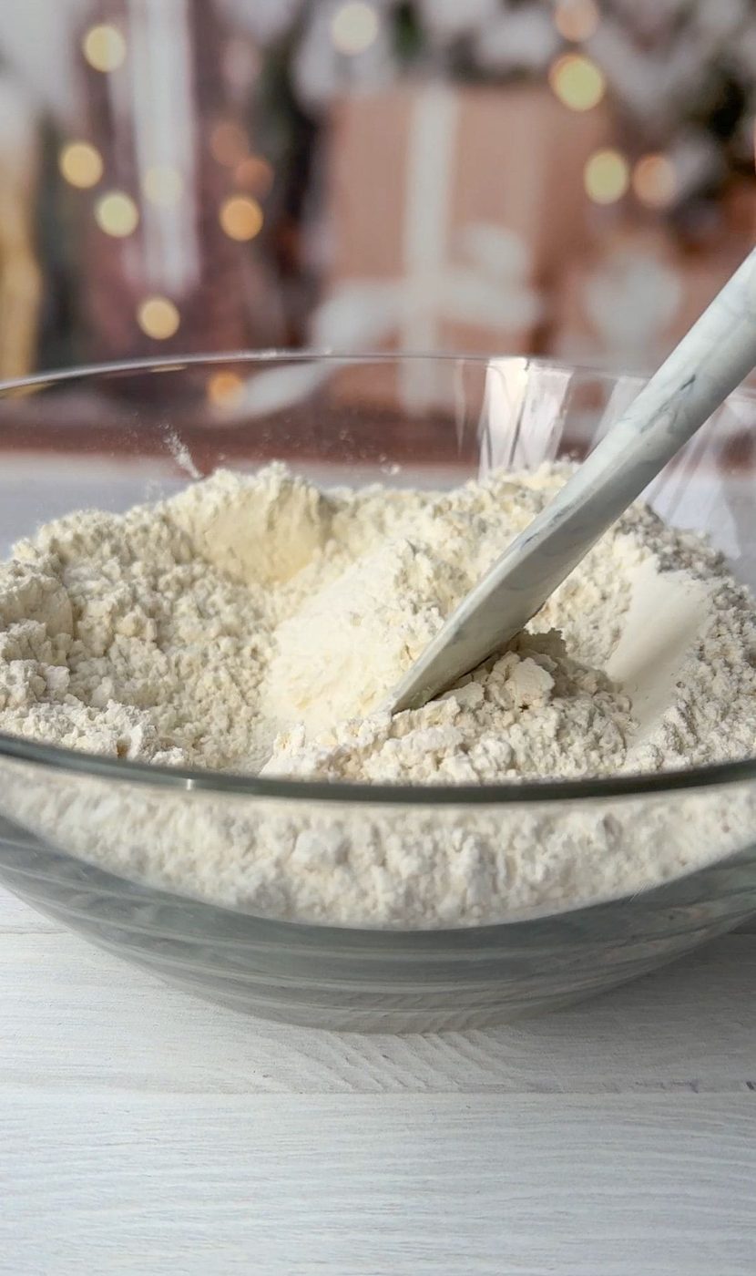 A glass bowl filled with flour and a white spatula resting in it. The background is softly blurred with warm, decorative lights and wrapped gifts, creating a festive atmosphere.