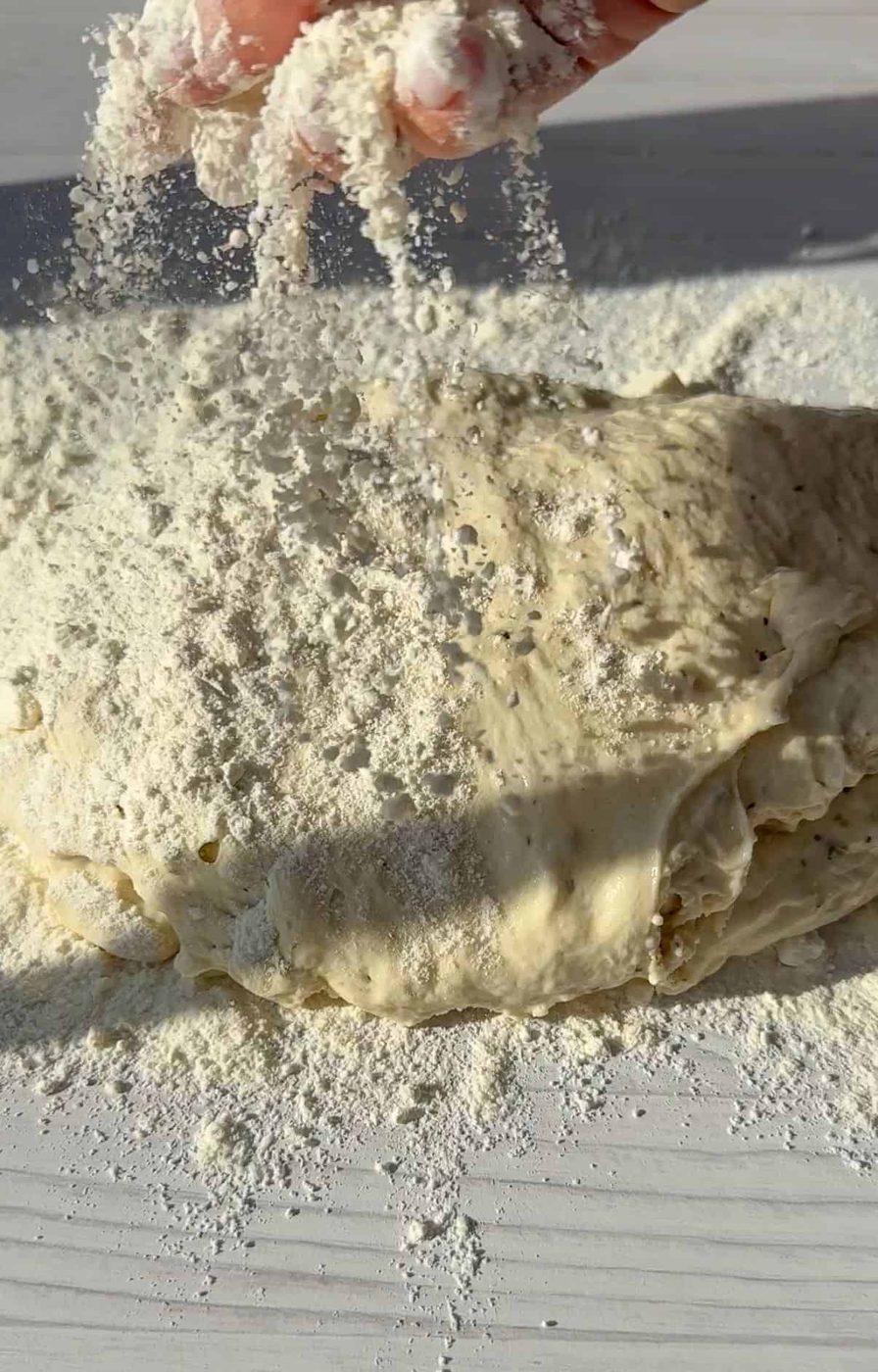 A hand sprinkling flour over a mound of no knead dough on a white surface, with sunlight casting soft shadows.