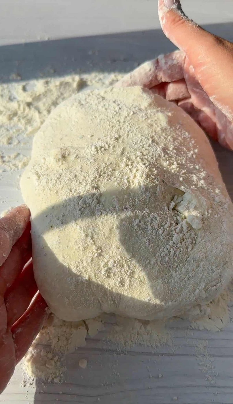 Hands kneading a dough ball covered in flour on a light-colored surface, with sunlight casting shadows.