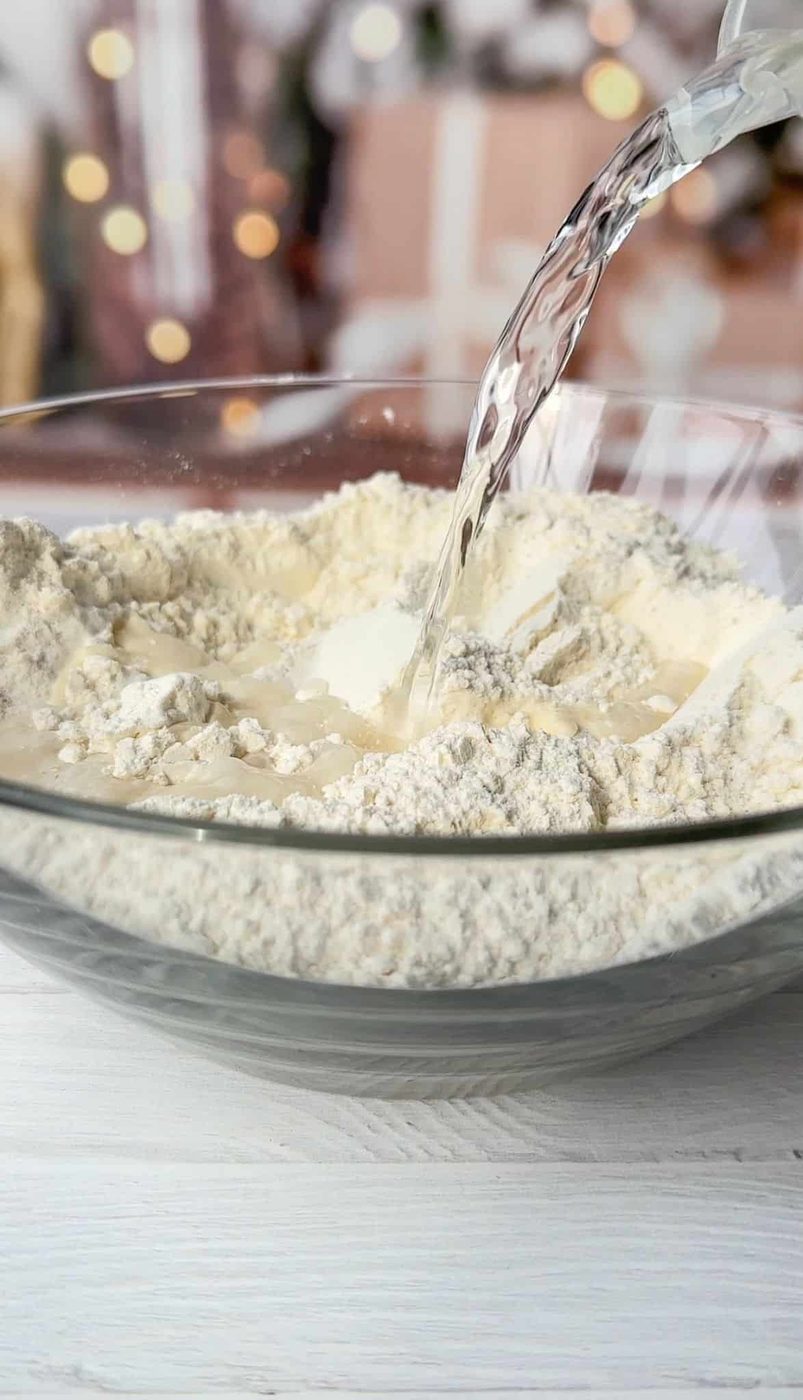 Clear liquid is being poured into a clear glass bowl filled with flour. In the background, there are blurred lights and indistinct shapes, creating a festive atmosphere.