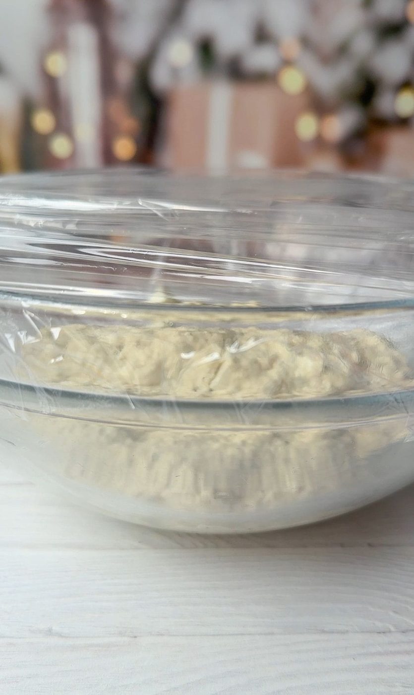 A glass bowl covered with plastic wrap, containing dough, sits on a white surface. Blurred background features soft lighting and hints of holiday decorations.