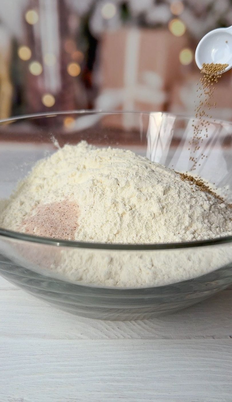 A clear glass bowl filled with flour and other dry ingredients. A spoon is pouring yeast into the bowl. There are blurred holiday decorations and wrapped presents in the background.
