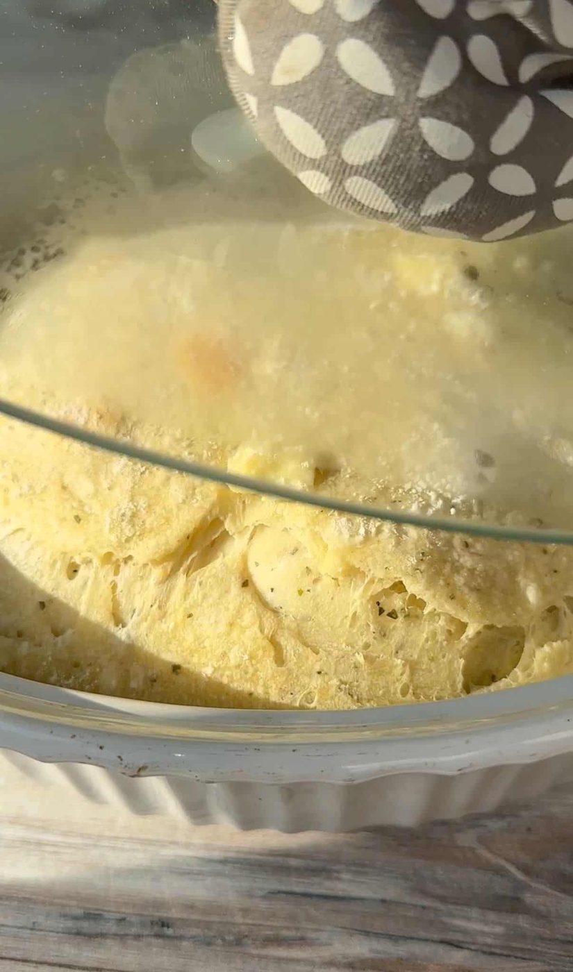 A close-up of a steamy No Knead Feta Bread being cooked under a glass lid. The dish appears to be a fluffy, baked meal with visible chunks, possibly cheese or vegetables. A hand holding a patterned oven mitt touches the lid.