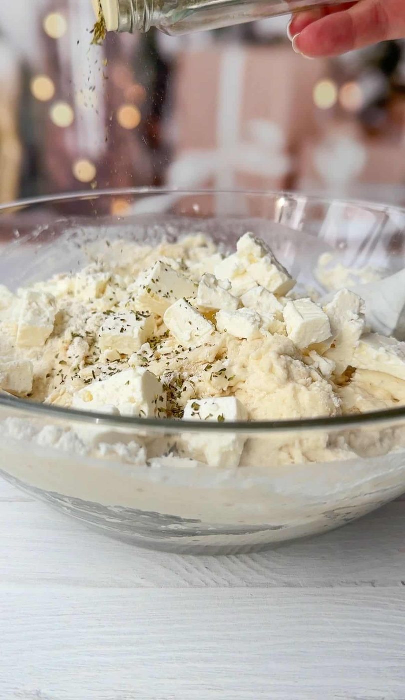 A clear glass bowl filled with chunks of feta cheese, creamy mixture, and herbs being sprinkled from a shaker above. The background is softly blurred, emphasizing the preparation of a dish on a white surface.