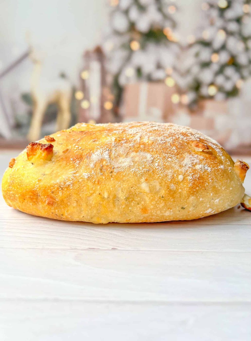 A freshly baked, golden-brown loaf of No Knead Feta Bread bread dusted with flour sits on a light wooden surface. The background is softly blurred with hints of festive decorations, including lights and greenery.