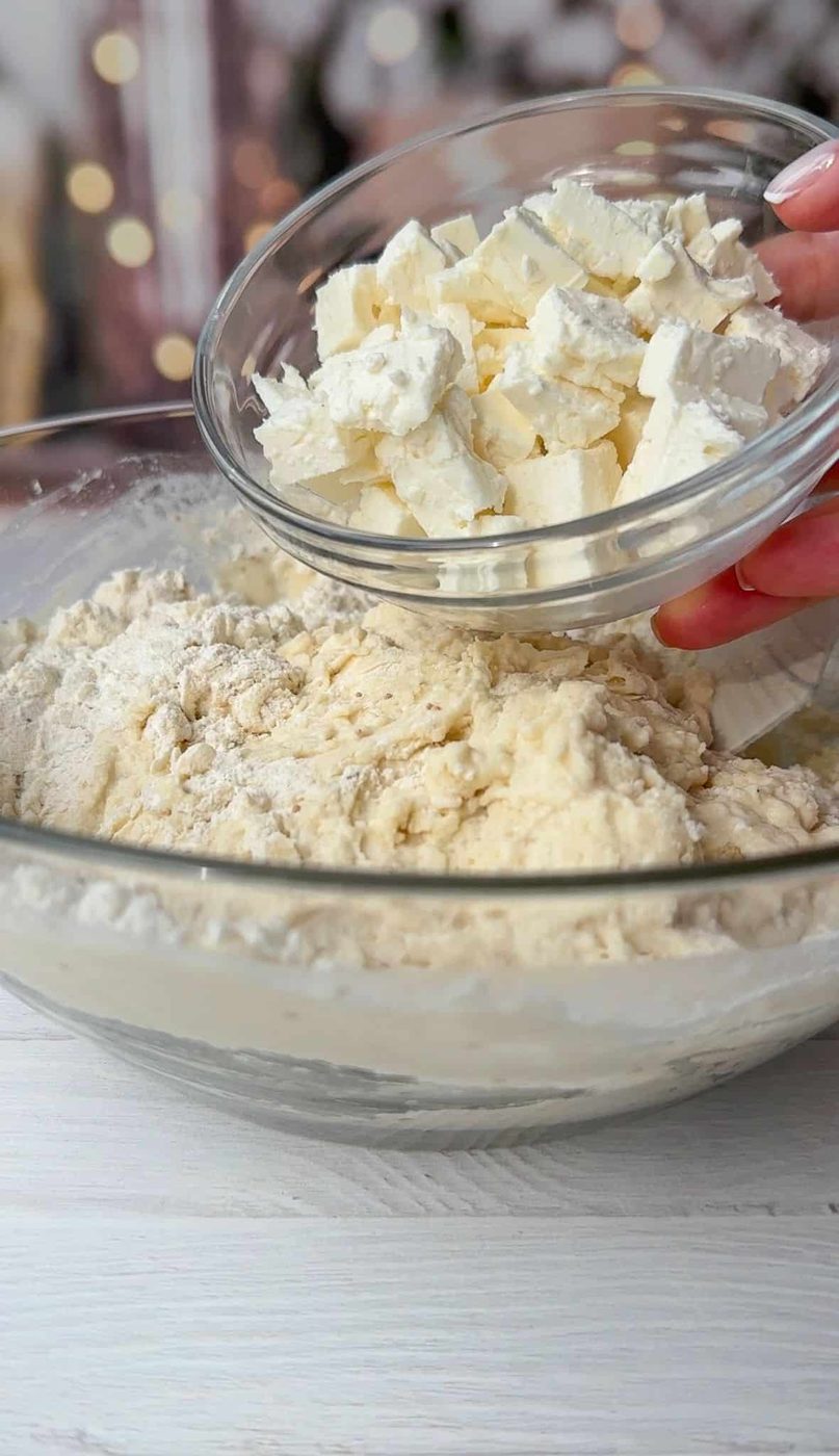 A hand holding a small bowl of cubed butter is adding it to a large mixing bowl filled with flour. The ingredients are on a white wooden surface, with blurred lights in the background.