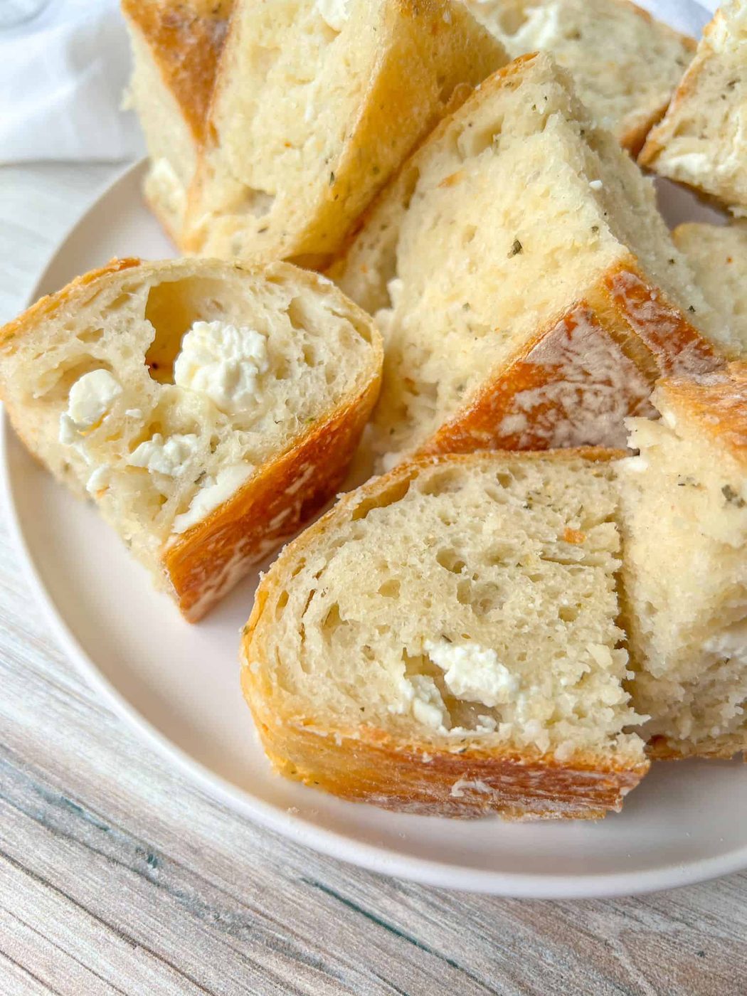 A plate filled with slices of crusty No Knead Feta Bread, featuring a golden-brown crust and soft, airy interior. The bread has visible pockets of cheese, adding texture and flavor. The plate is set on a light wooden surface.