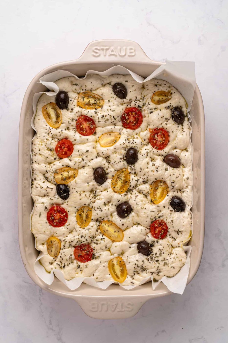 A rectangular dish of focaccia dough topped with cherry tomatoes, olives, and herbs. The dough is placed on parchment paper, ready for baking, in a cream-colored ceramic dish on a white marble surface.