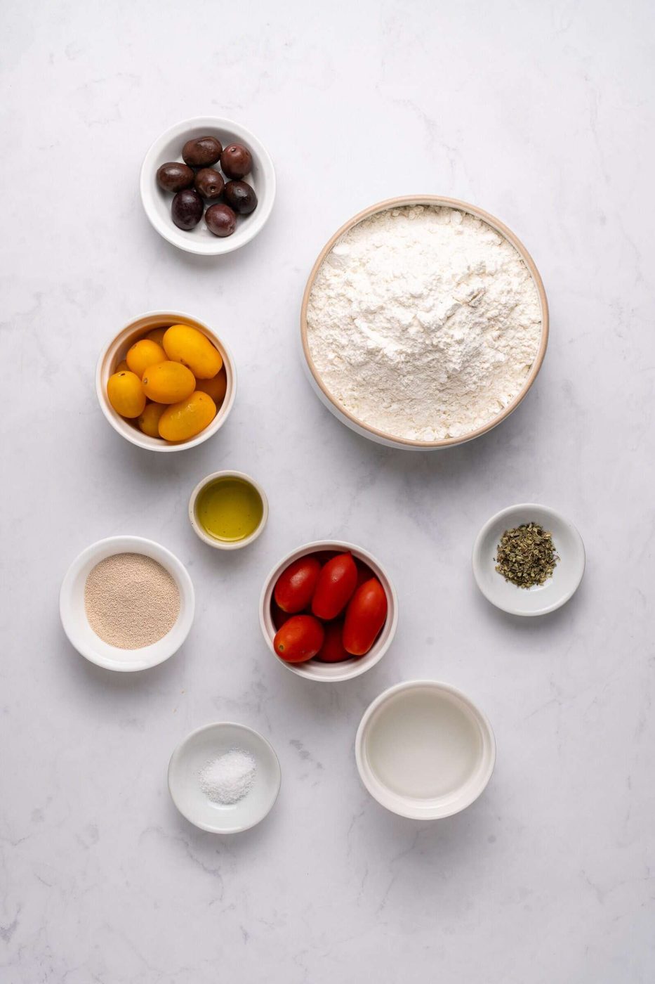Bowls with flour, yellow and red tomatoes, olives, olive oil, yeast, oregano, salt, and water on a white surface.