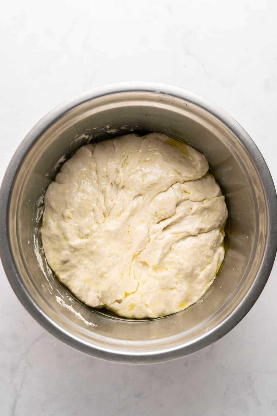 A bowl of unbaked dough rests in a metal container. The dough is slightly shiny and textured, suggesting olive oil on its surface. The background is a light, marbled countertop.