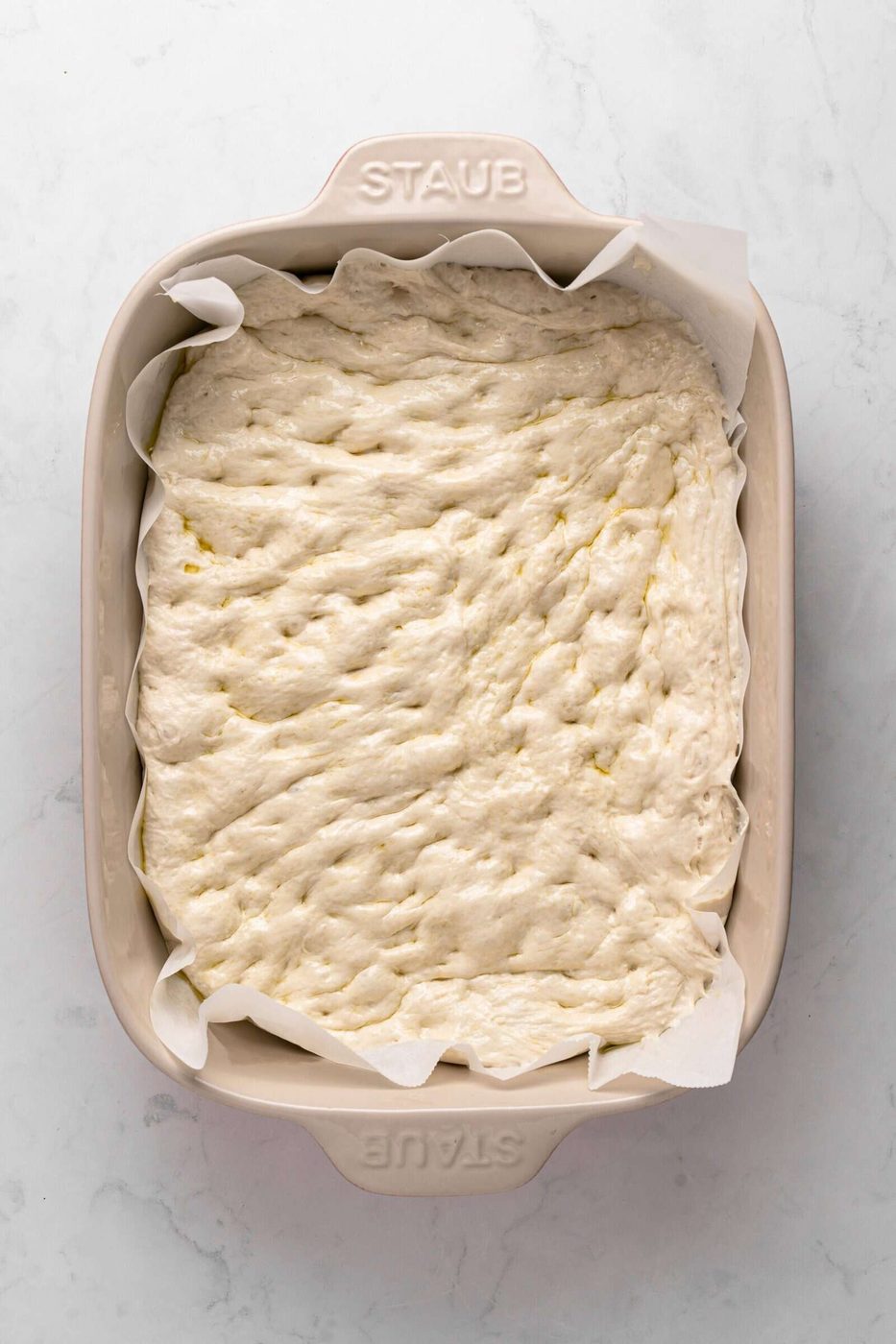 A rectangular baking dish lined with parchment paper contains raw focaccia dough, visibly dimpled and ready to be baked. The dish is light-colored and labeled "STAUB" on the handles. The background is a light, textured surface.