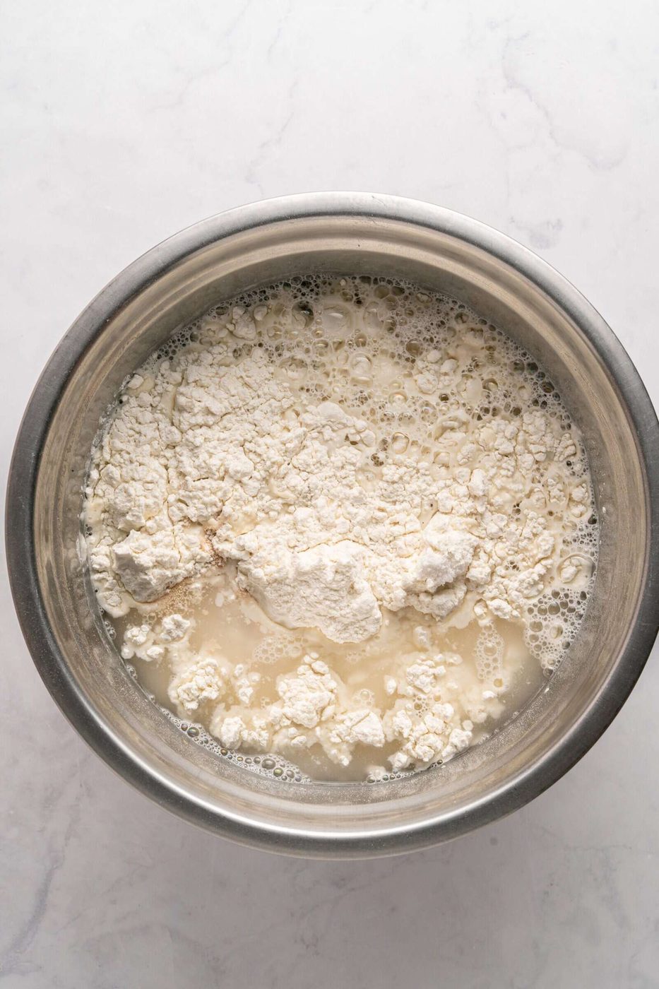 A stainless steel bowl containing flour, water, and other ingredients, possibly yeast, is ready for mixing on a light marble countertop.