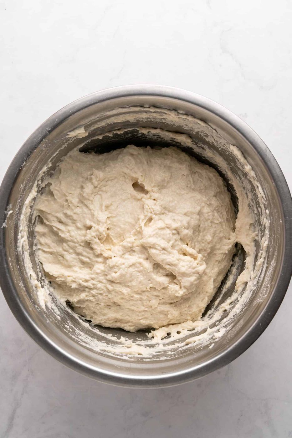 A stainless steel bowl filled with smooth, mixed bread dough sits on a white marbled surface.