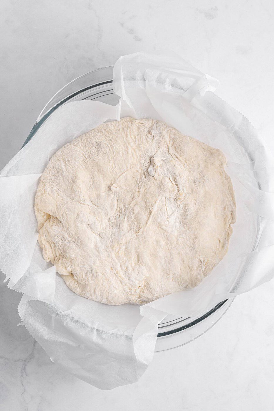 A bowl lined with parchment paper holds a round, lightly floured dough on a white marble surface. The dough appears soft and slightly dimpled, ready for baking or further preparation.
