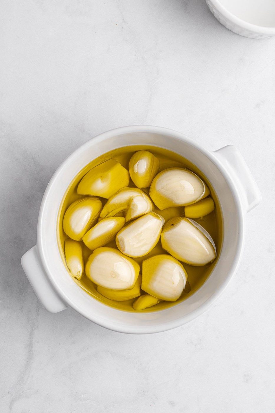 A white bowl filled with several peeled garlic cloves submerged in golden olive oil, placed on a light marble surface.