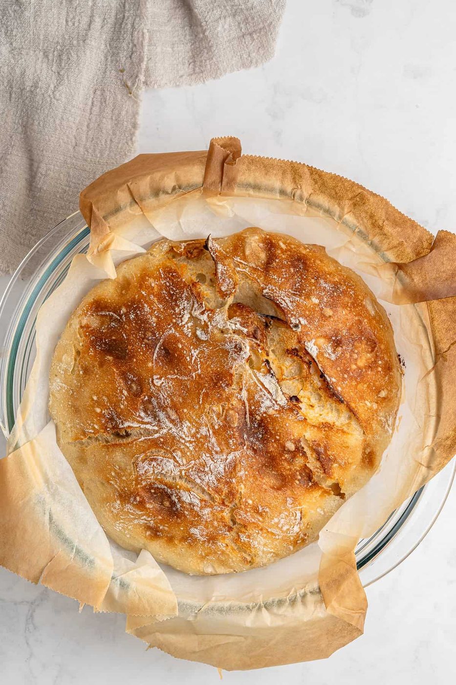 A freshly baked round loaf of crusty No Knead Garlic Bread on parchment paper in a glass dish. The bread has a golden-brown crust with a dusting of flour. A beige cloth is partially visible in the top left corner on a light-colored surface.