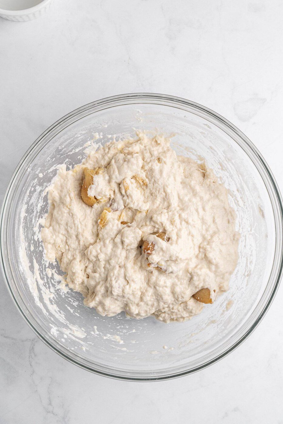 A glass bowl filled with a thick, creamy bread dough mixture on a white marble countertop.