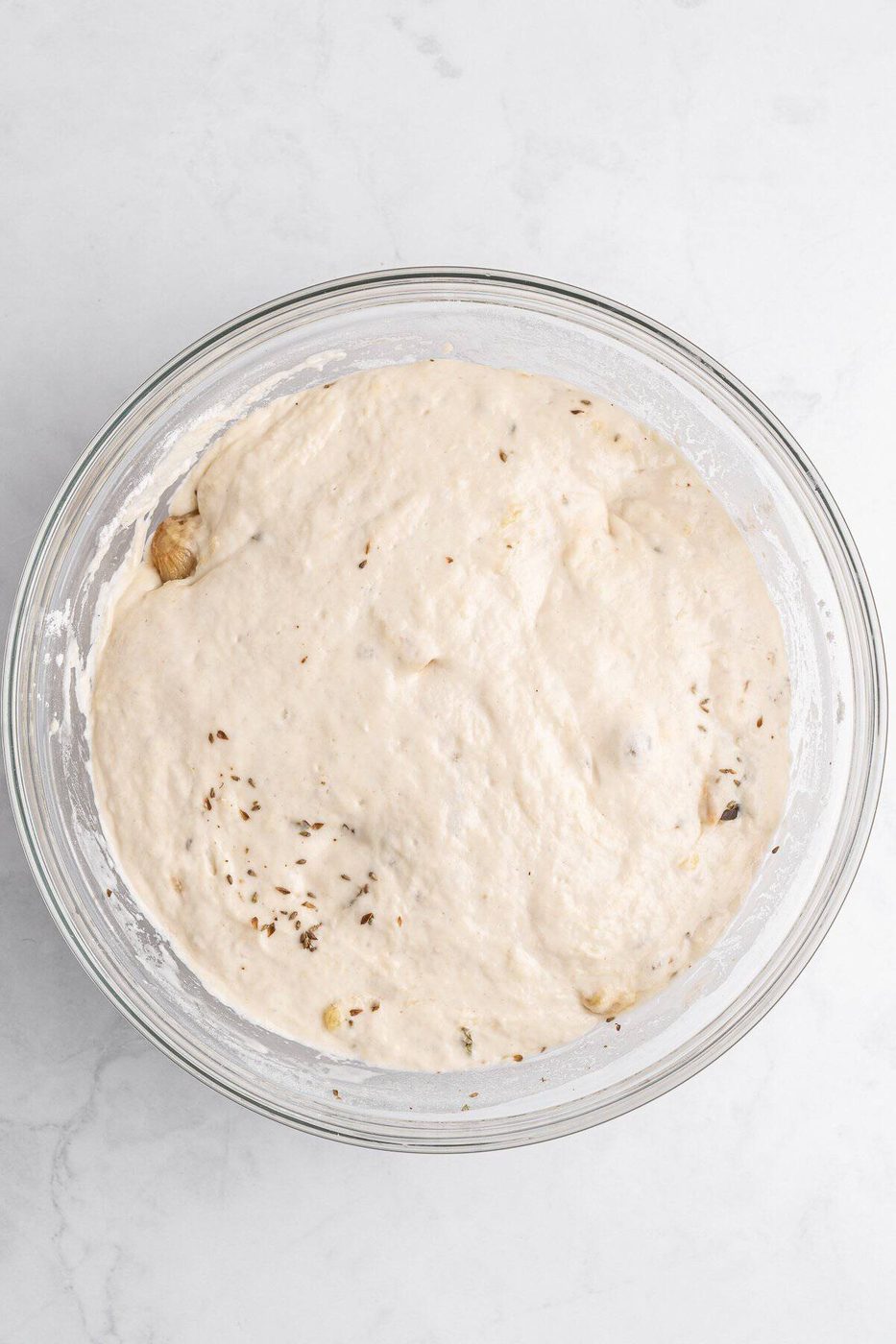 A glass bowl filled with pizza dough rising, with visible herbs and small ingredients mixed in. The bowl is placed on a white marble surface.