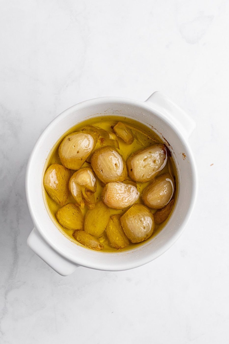 A white bowl filled with garlic cloves submerged in golden olive oil. The bowl is set against a light marble surface. The garlic appears roasted, with a soft texture and light browning.
