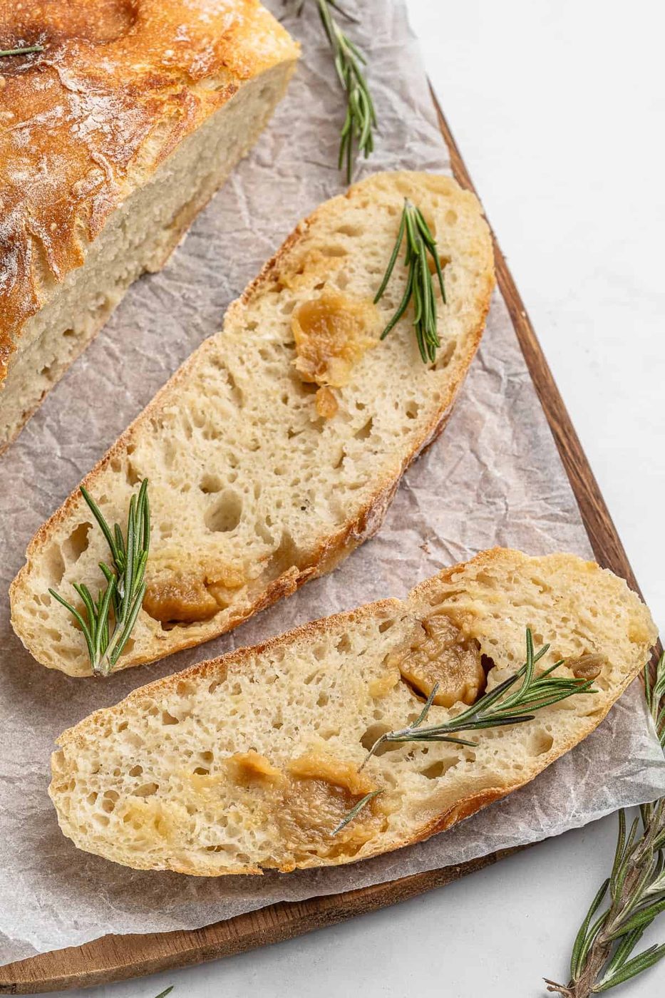 Two slices of crusty No Knead Garlic Bread topped with roasted garlic and sprigs of rosemary rest on parchment paper on a wooden cutting board. A portion of a loaf is visible in the background. Sprigs of rosemary are scattered around.