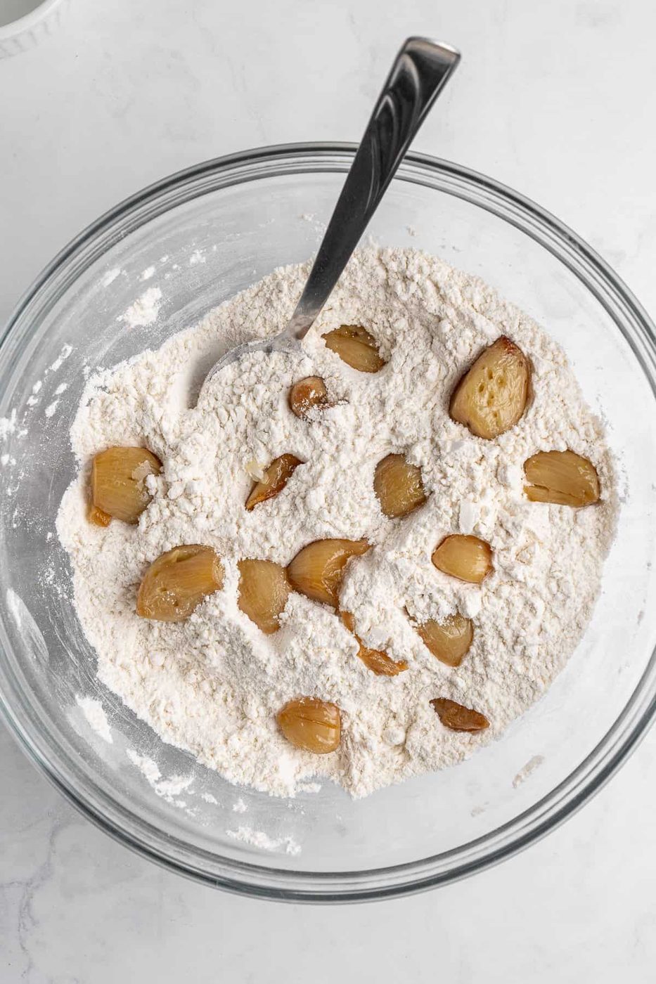 A glass bowl filled with flour mixed with roasted garlic cloves. A spoon is partially submerged in the flour mixture, and the bowl is set on a light-colored surface.