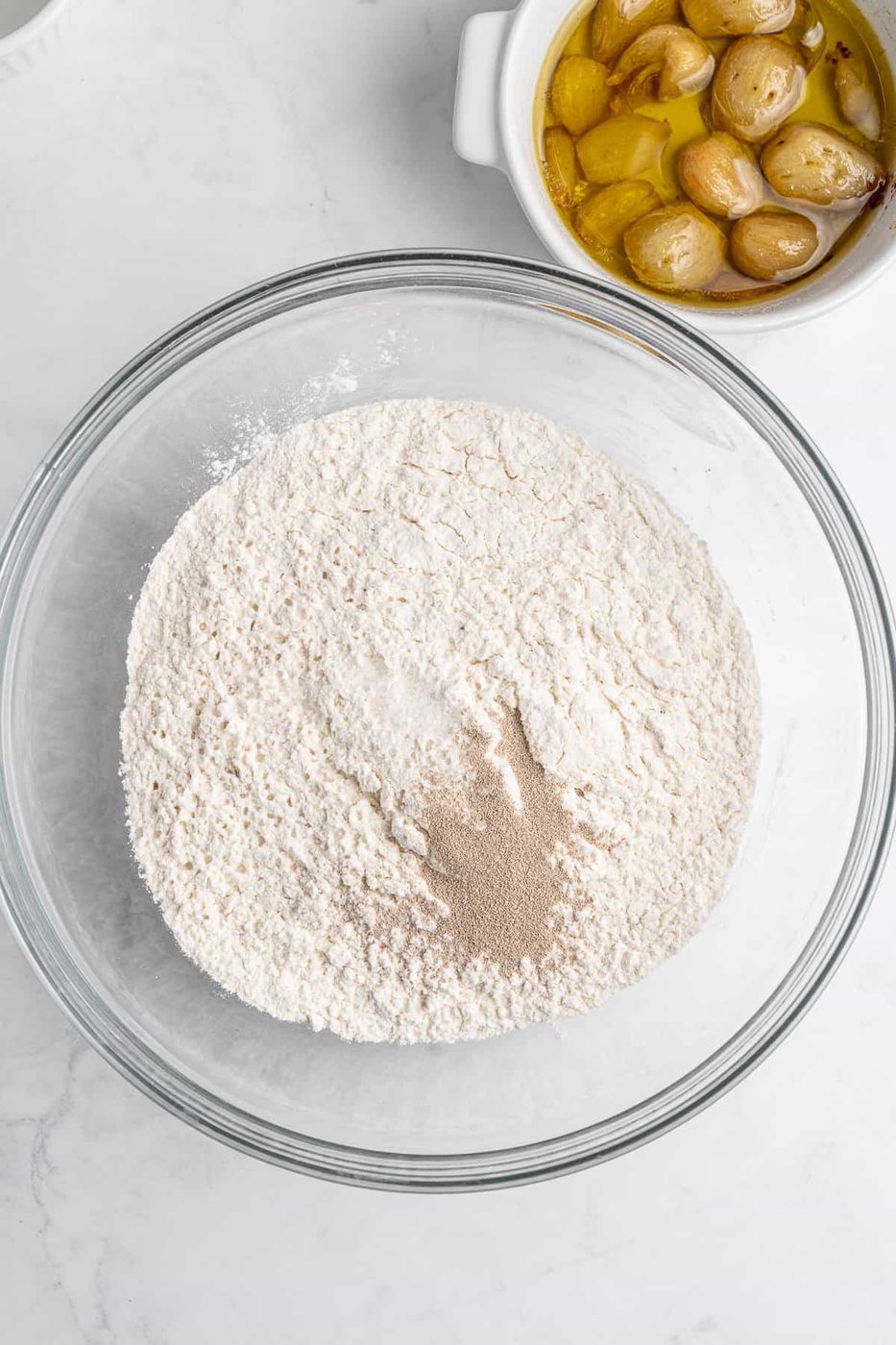 A glass bowl containing a mixture of flour and yeast sits on a marble surface. Next to it is a bowl of roasted garlic cloves in olive oil.