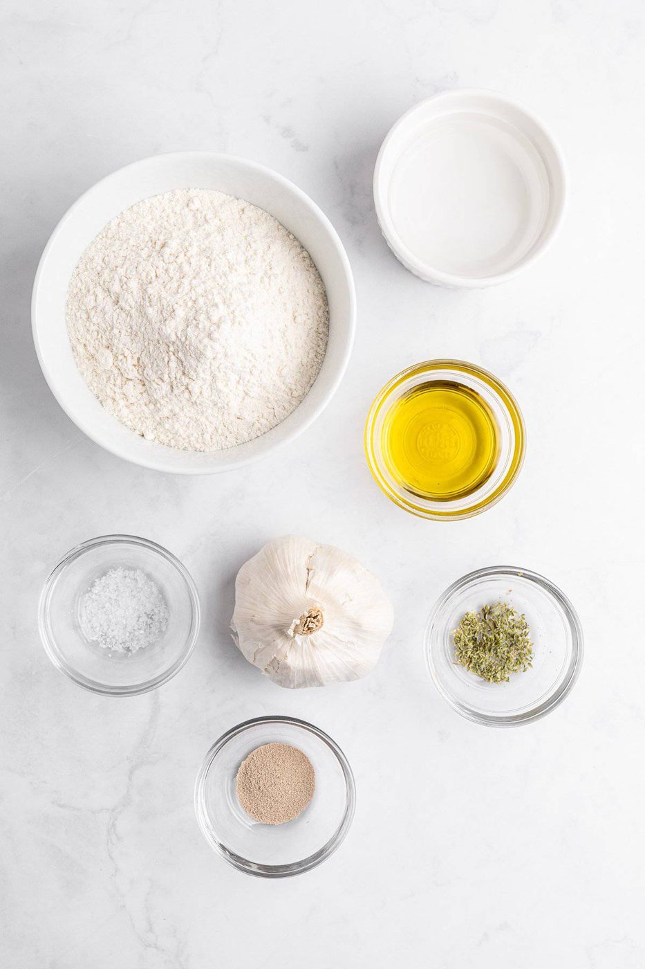 Ingredients on a white surface: a bowl of flour, a small bowl of water, a bowl of olive oil, a garlic bulb, a dish of dried herbs, a dish of salt, and a dish of yeast.