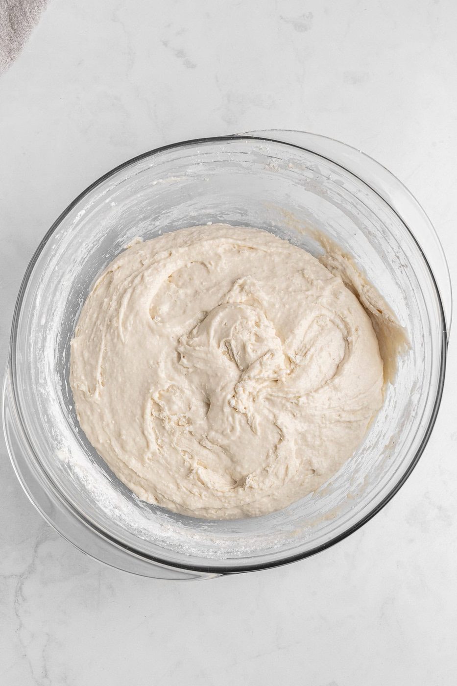 A glass bowl filled with smooth, creamy batter sits on a white marble surface. The mixture is light beige in color, with a slightly glossy texture, indicating it's well-mixed and ready for baking.