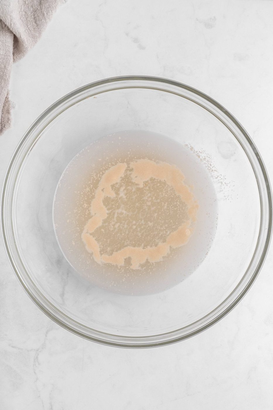 A transparent glass bowl sits on a marble countertop, containing water with dissolved yeast. The yeast forms an uneven, frothy beige layer on the surface, with some granules scattered throughout. A beige cloth is partially visible in the top left corner.