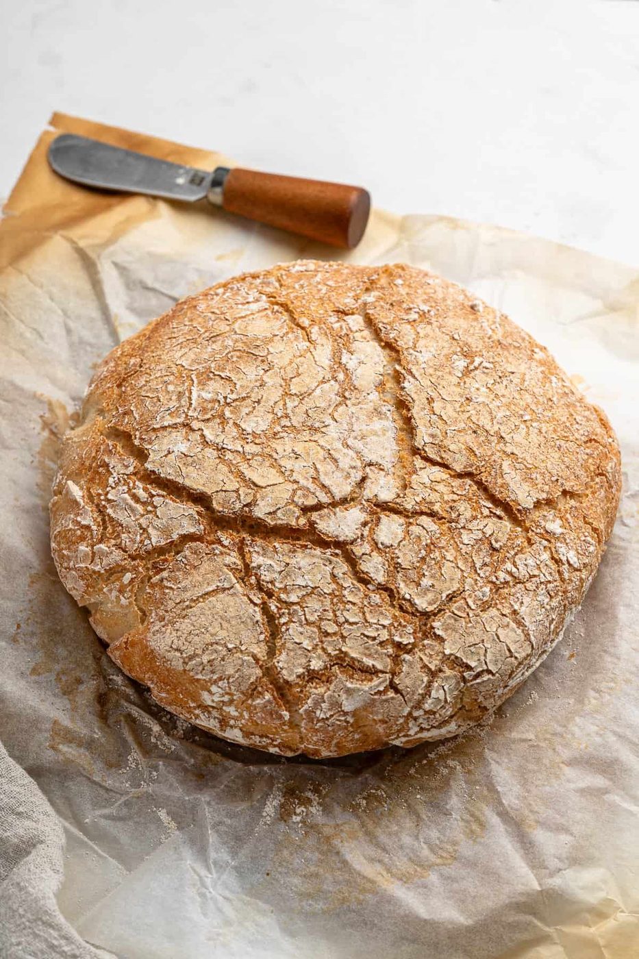 A round loaf of rustic, crusty bread with a cracked surface sits on parchment paper. Beside it is a butter knife with a wooden handle, placed on a piece of brown paper. The background is a light, textured surface.