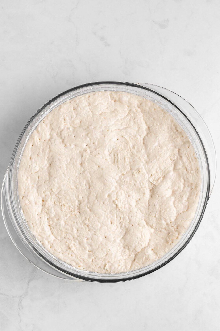 A glass bowl filled with risen bread dough, resting on a light gray marble surface. The dough has a smooth and slightly textured appearance, indicating it has been proofed and is ready for the next step in baking.