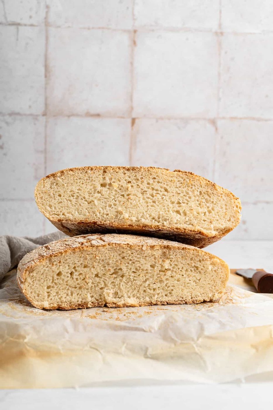 Two halves of a rustic loaf of bread are stacked on parchment paper. The texture of the bread's crumb is visible, and the crust has a lightly toasted appearance. A knife handle is partially visible on the right side. The background is tiled.