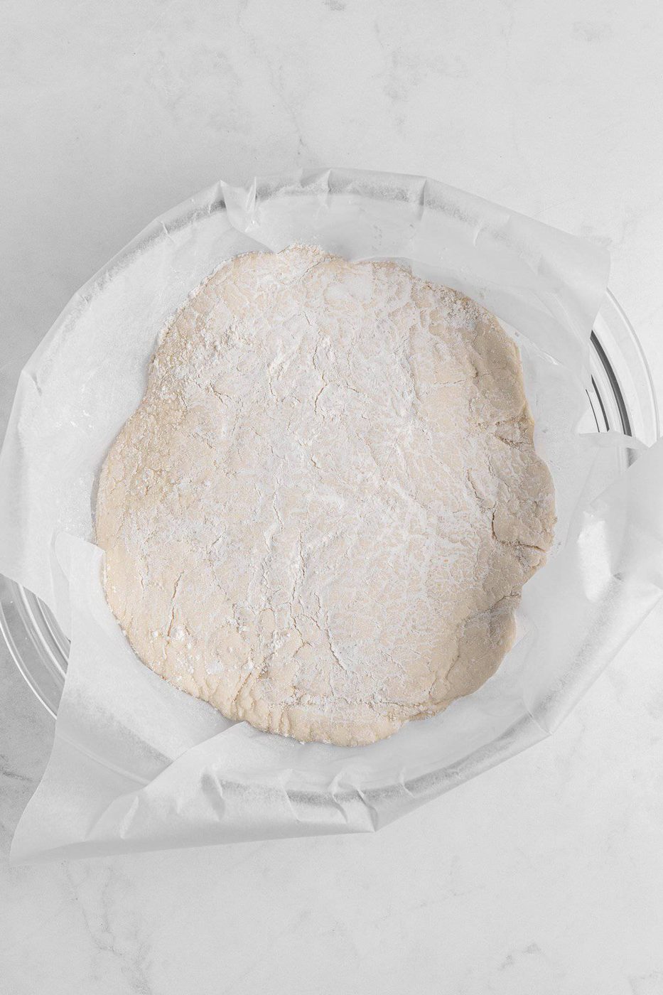 A ball of dough rolled out flat on parchment paper in a glass bowl. The dough is lightly dusted with flour and sits on a white marble surface.