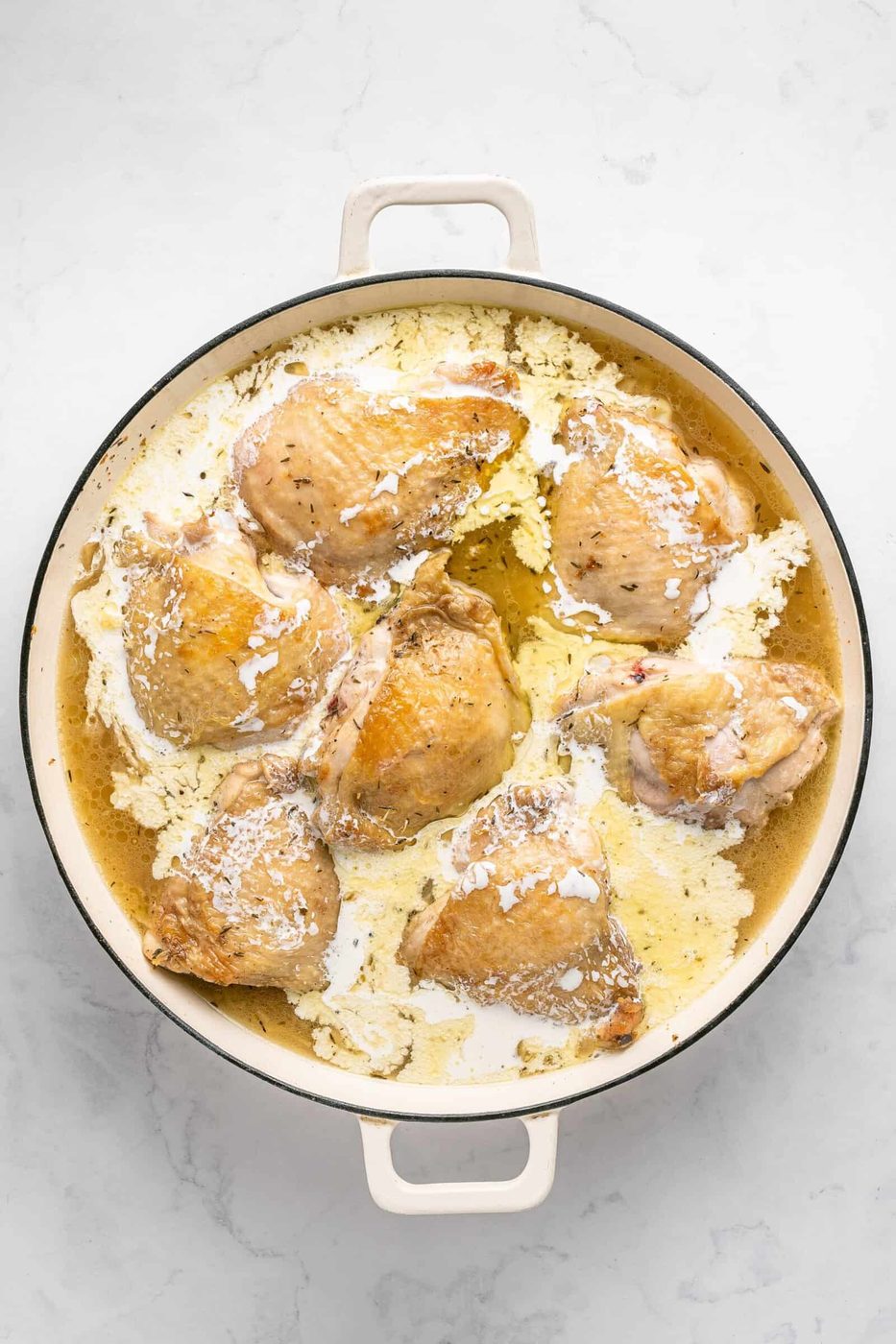 A round white Dutch oven filled with several browned chicken thighs simmering in a creamy sauce on a light marble surface—a comforting one pan chicken dinner perfect for any night.