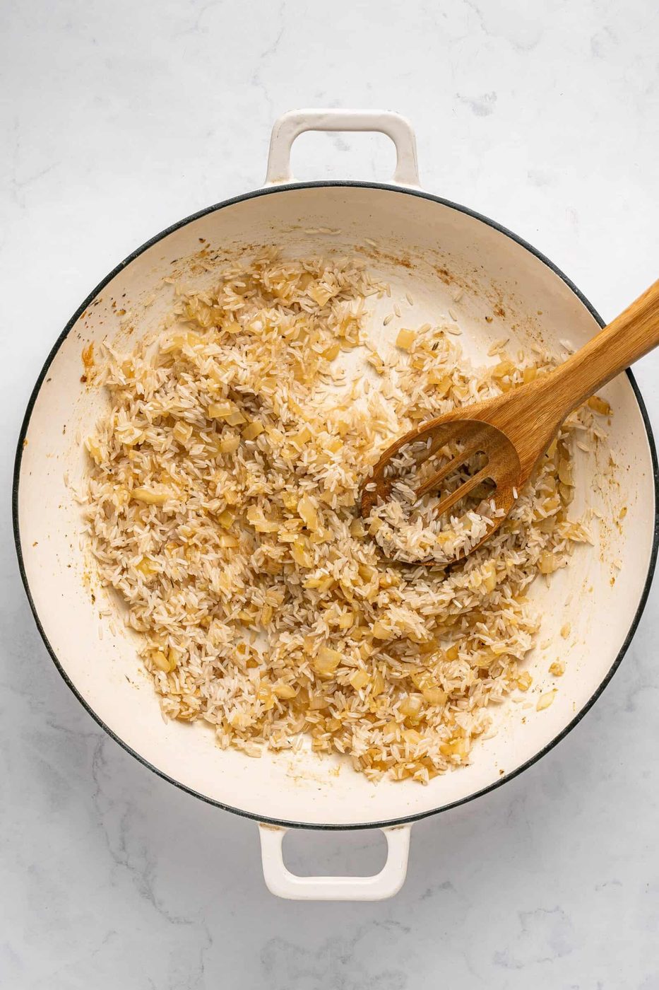 A white pot filled with sautéed onions and uncooked rice for a delicious One Pan Chicken Dinner, being stirred with a wooden spoon on a light-colored countertop.