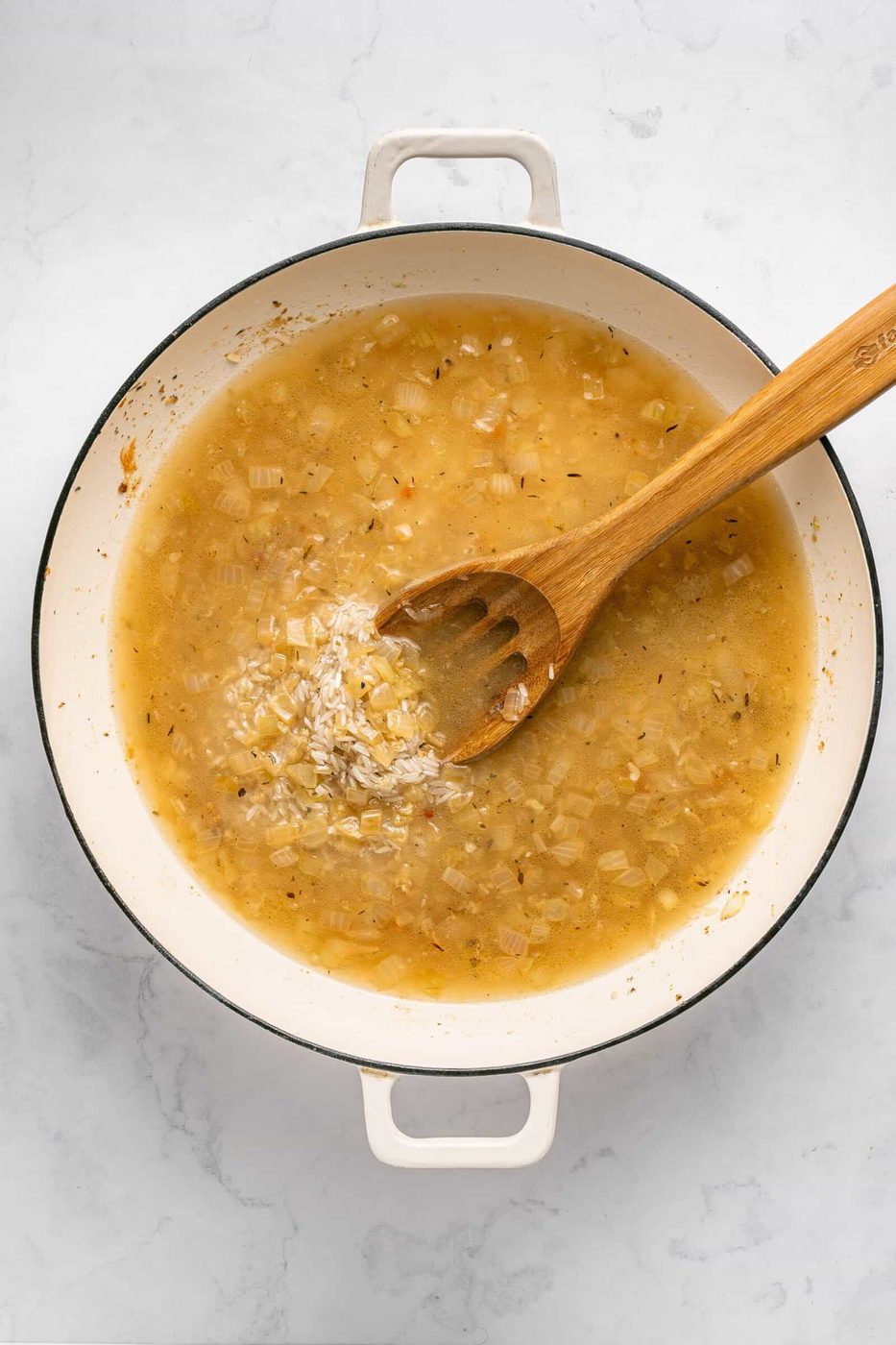 A white pot containing a mixture of chopped onions, broth, and seasonings being stirred with a wooden spoon. Uncooked rice has just been added—perfect for starting a flavorful One Pan Chicken Dinner on a light-colored countertop.