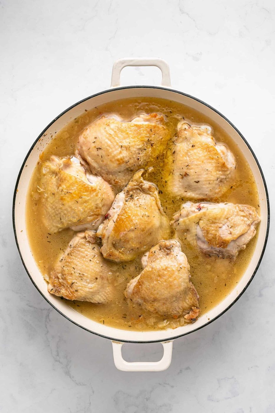 A round white Dutch oven filled with seven browned chicken thighs simmering in broth on a light marble surface—a perfect One Pan Chicken Dinner for effortless, delicious meals.