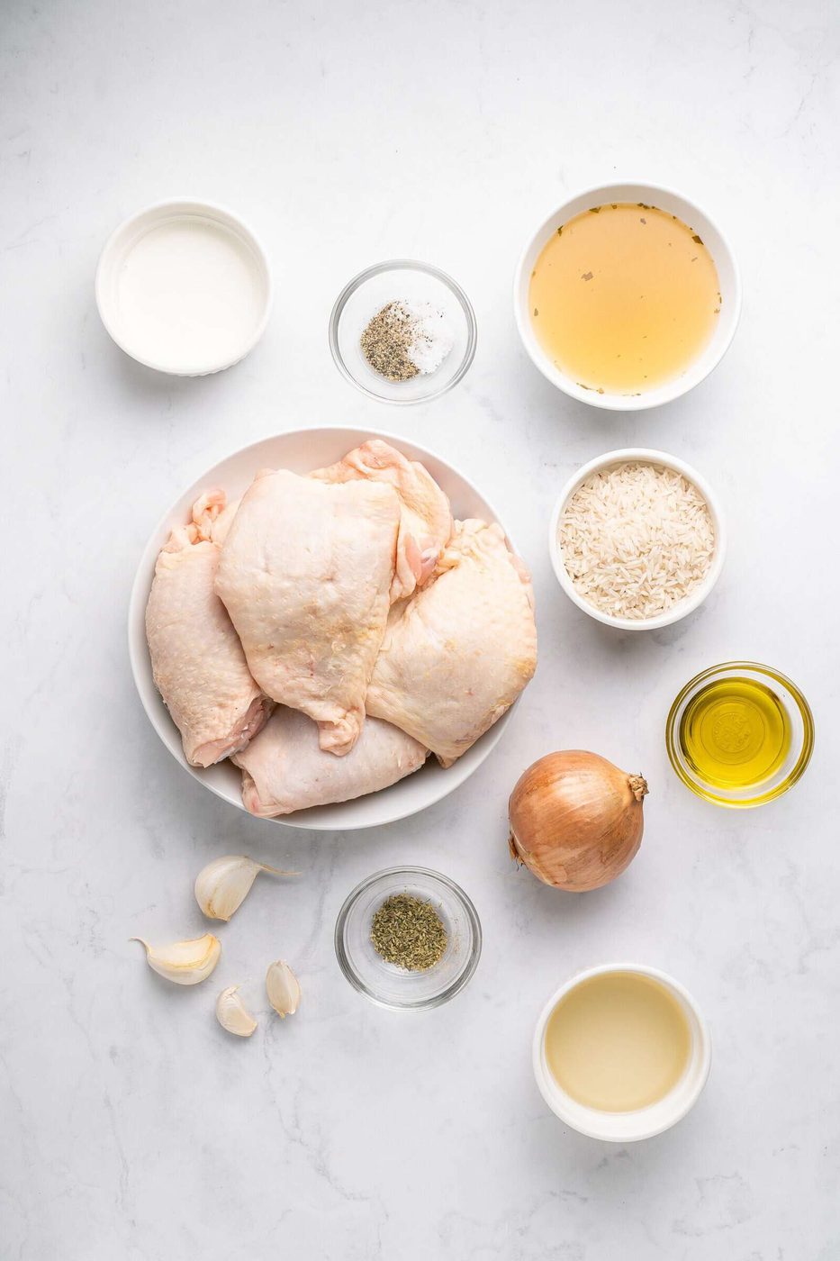 A top view of a cooking setup with raw chicken thighs on a plate, surrounded by bowls of salt, pepper, olive oil, broth, rice, milk, garlic cloves, and an onion on a white marble surface.