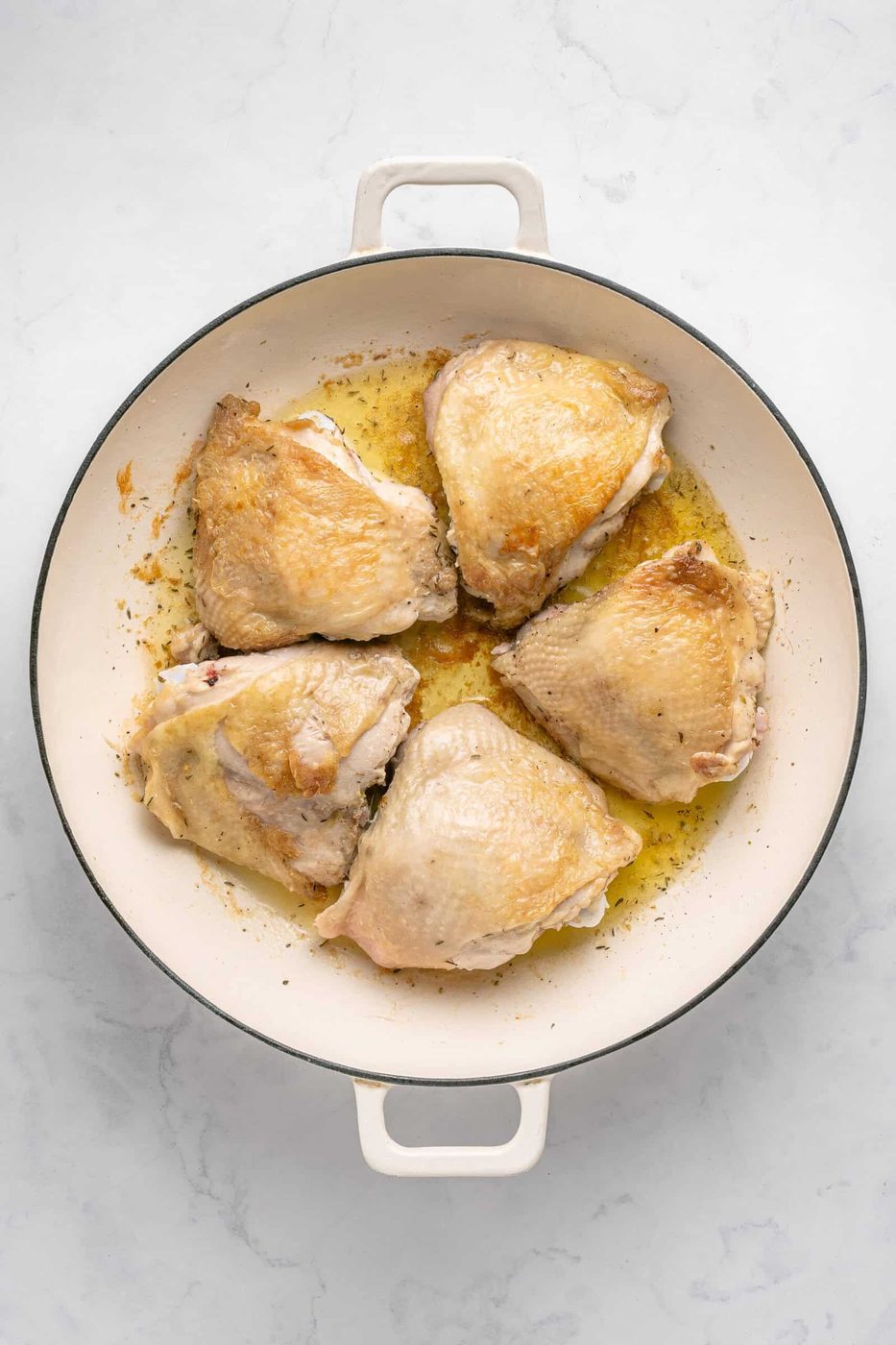 Five browned chicken thighs sizzle in melted butter or oil in a white enameled skillet, creating the perfect One Pan Chicken Dinner, seen from above on a light marble surface.