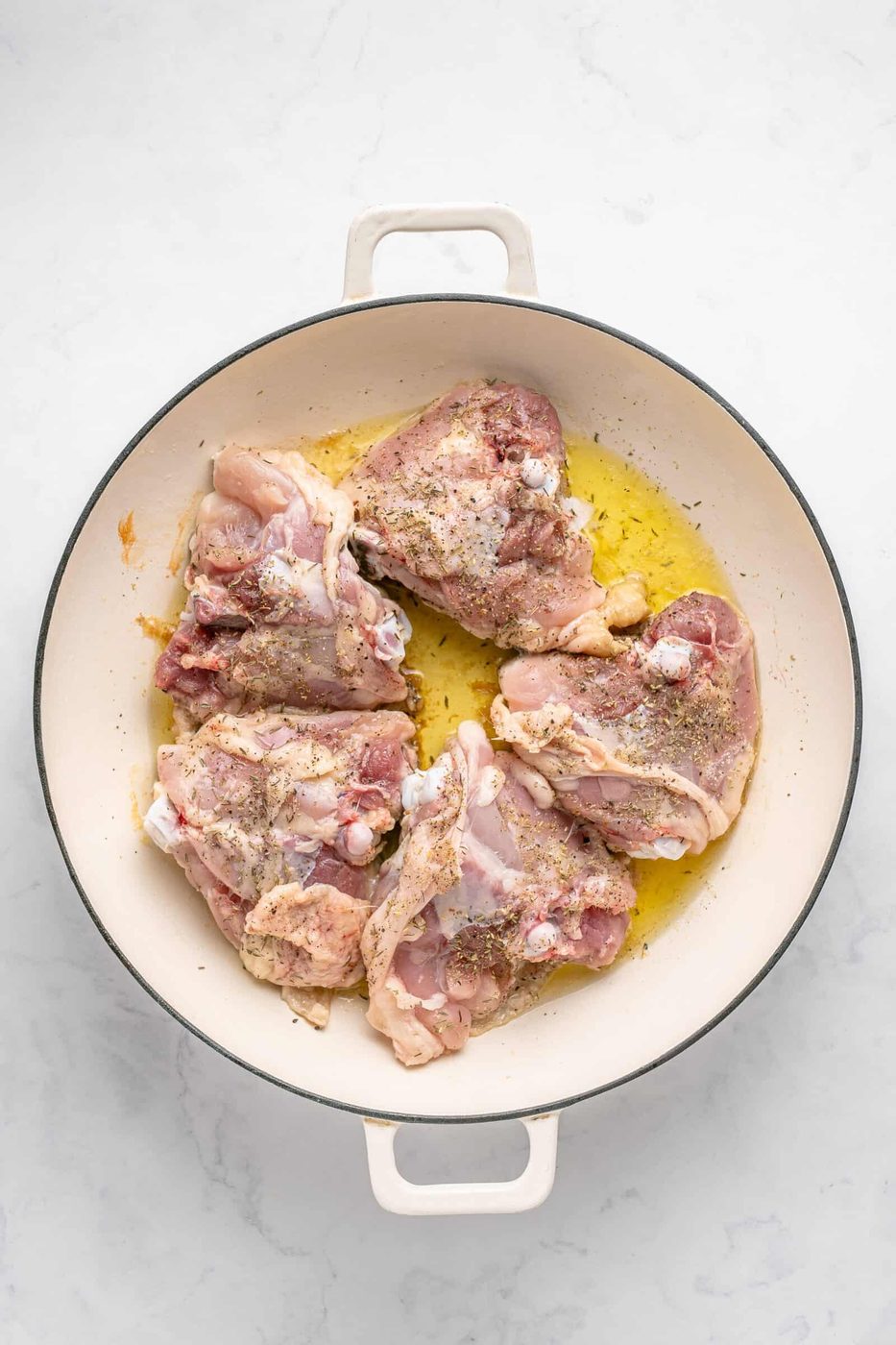 Raw chicken pieces seasoned with herbs and spices lie in a white cast iron skillet with a light coating of oil. The background is a marble-like surface, highlighting the preparation of the chicken for cooking.