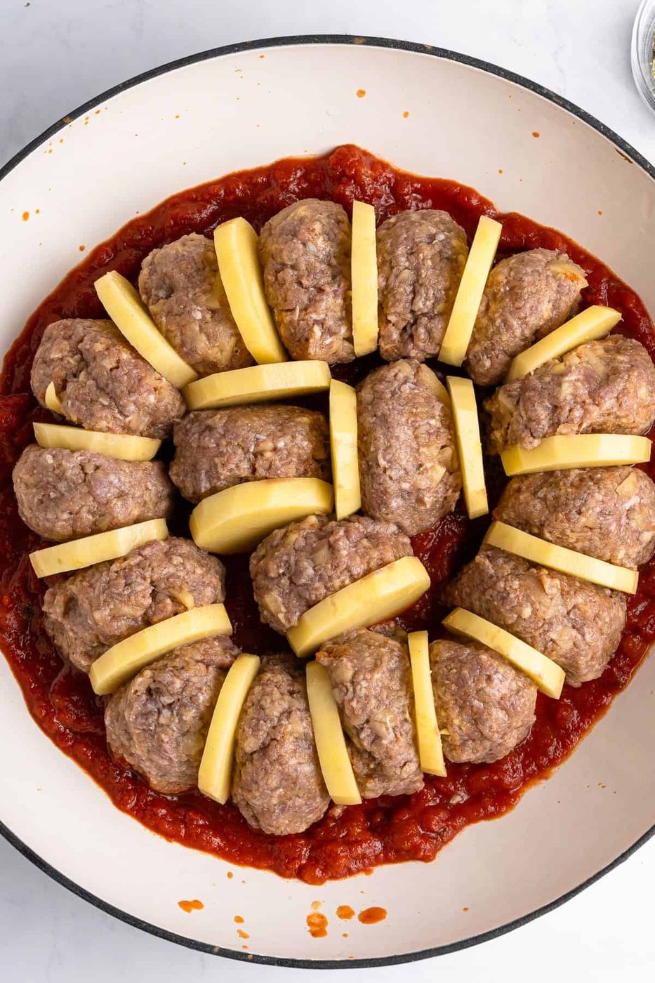 A round dish featuring a ring of uncooked meatloaf slices interspersed with potato slices, all placed over a thick layer of tomato sauce. The ingredients are neatly arranged, ready for cooking.