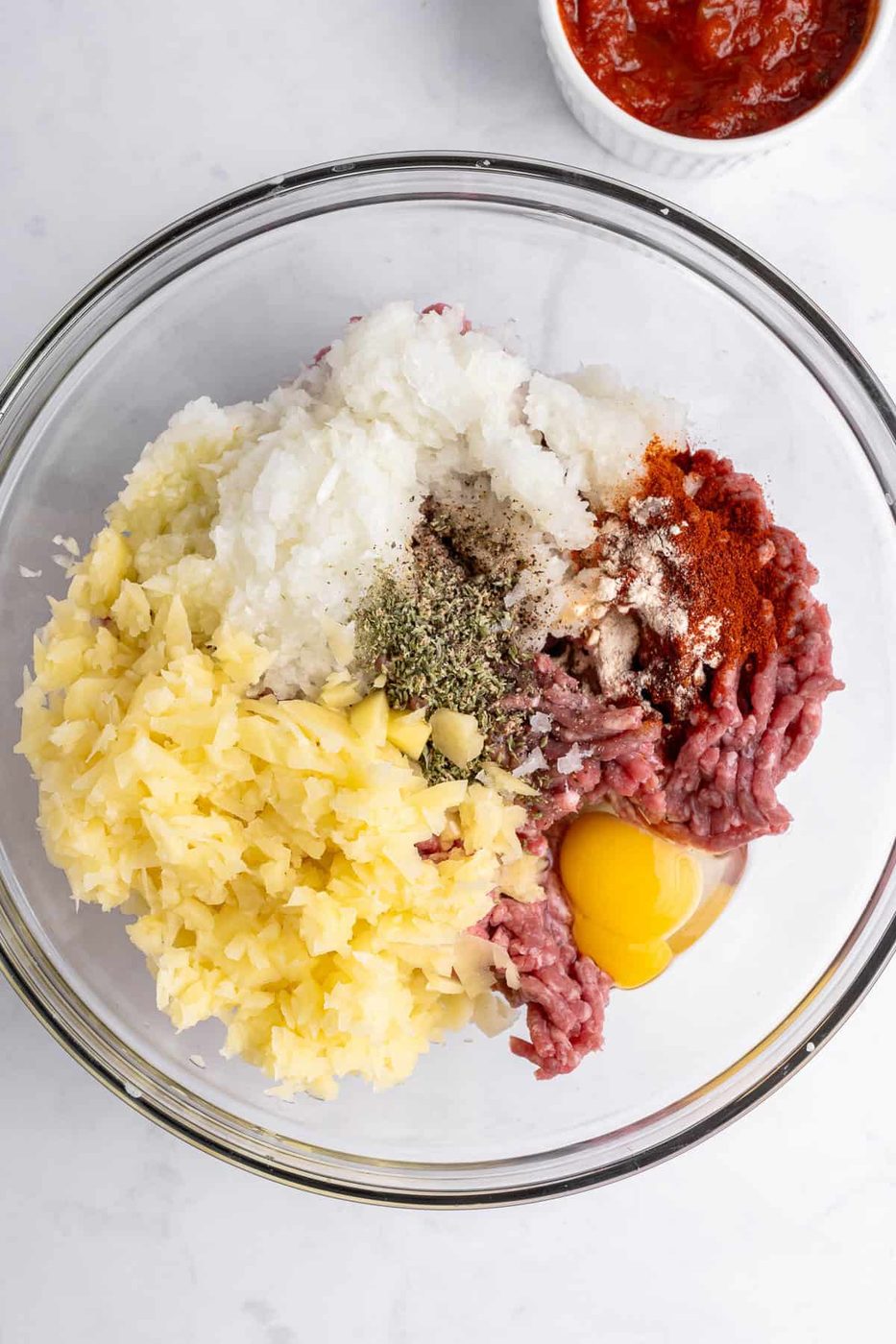 A glass bowl containing raw minced meat, chopped onions, potatoes, spices, and an egg yolk. A small bowl of red sauce is partially visible in the background on a white marble surface.